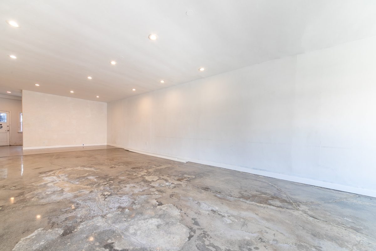 Vacant main room looking along the long white side wall, with polished concrete, recessed lighting, and a rear opening at the far left.