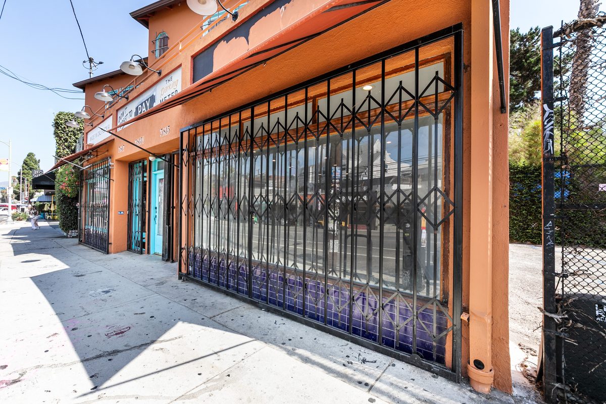 Oblique sidewalk view of the 3815 display window behind a folding security gate, with purple tile at the base and the neighboring 3817 bay in the distance.